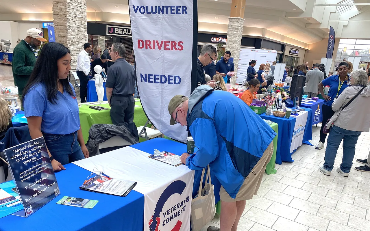 A man signing up to become a volunteer driver at an outreach table beneath a Volunteer Drivers Needed banner.