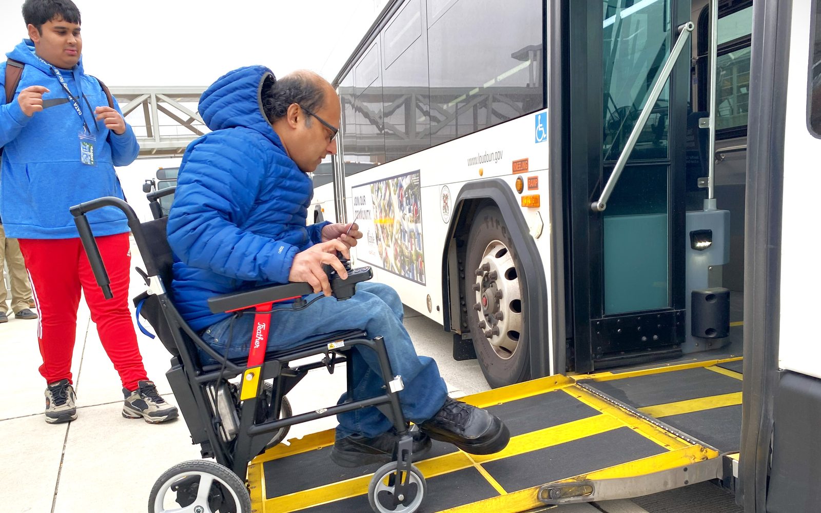 A man in a power wheelchair steers up the fold-out ramp of a Loudoun County Transit bus while a fellow Transit Treks participant watches from the platform.