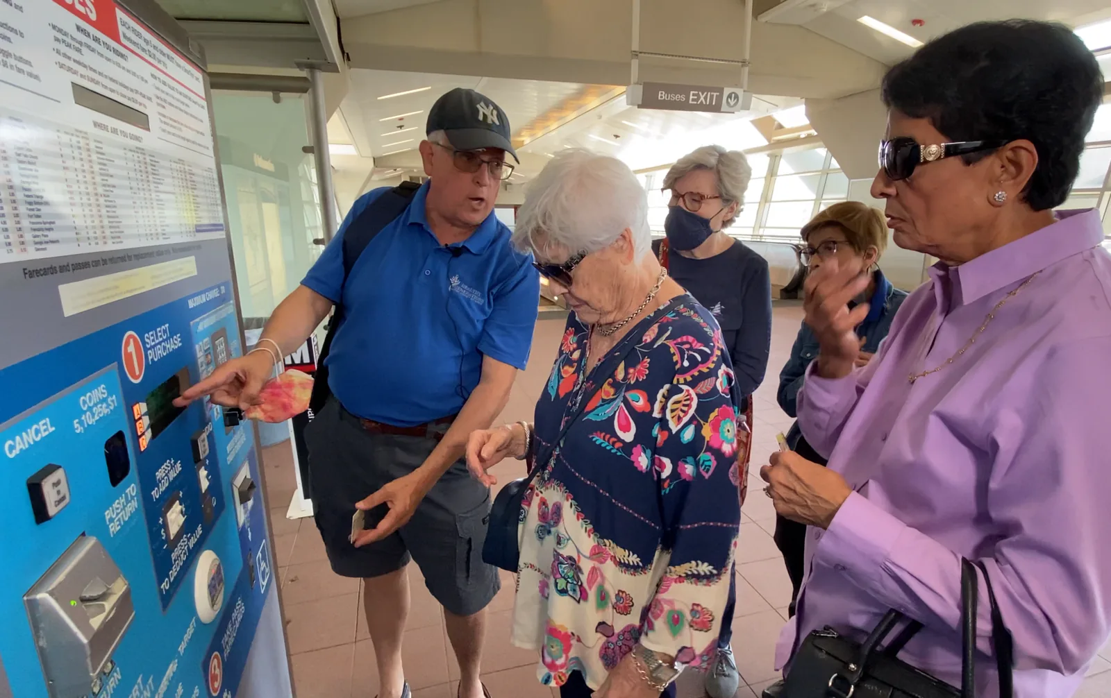 A DATA staff member points out the buttons on a Metro fare vending machine while four older adults watch, with the fare chart posted on the wall beside them.